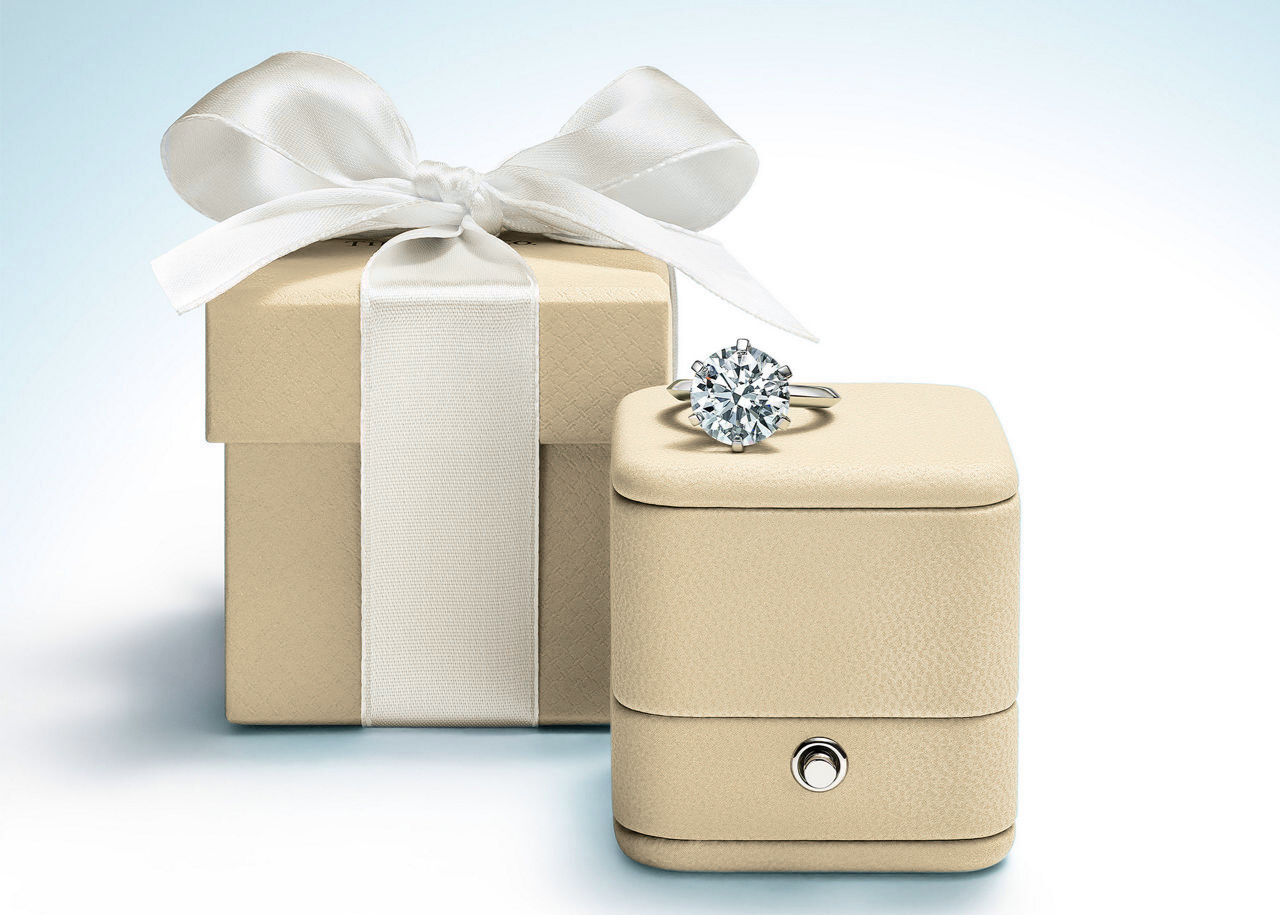 Beige jewelry box with a diamond ring on top and a matching gift box with a white ribbon.