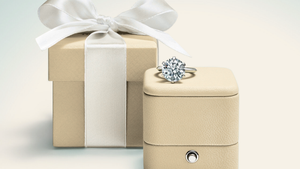 Beige jewelry box with a diamond ring on top and a matching gift box with a white ribbon.