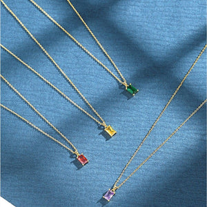 A collection of baguette cut birthstone pendant necklaces in various colors, set in gold plating, displayed on a blue background.