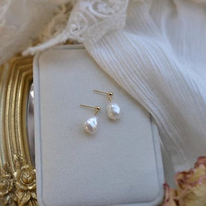 A set of three baroque pearl dangle drop earrings with gold plating, displayed on a white surface with a dried flower and part of a white garment visible in the background.