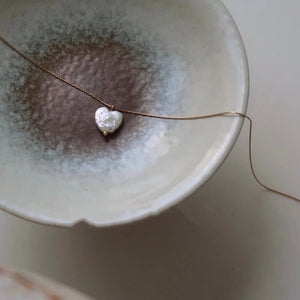 Heart-shaped pearl pendant on a thin gold chain over a ceramic dish.