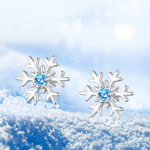 A pair of sterling silver snowflake stud earrings with blue cubic zirconia centers, displayed against a snowy backdrop.