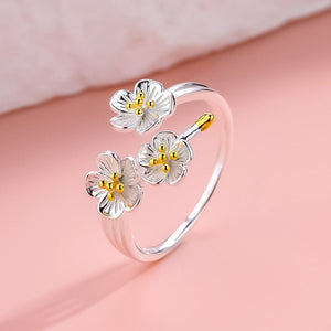 An image featuring two sterling silver rings with cherry blossom flower designs, one open band and one bypass ring, displayed on a pink background.