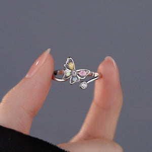 Hand holding a butterfly-shaped ring with colorful gemstones on a gray background