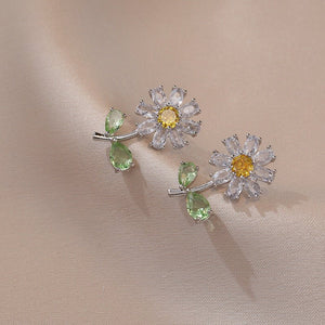A pair of gold-plated flower stud earrings with a central yellow gemstone and green leaf accents on a silky background.