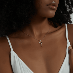 A close-up of a model wearing a delicate gold chain necklace with a small gold cross pendant featuring a dark blue stone, styled with a white camisole.