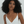 A woman with voluminous curly black hair wearing a white spaghetti-strap top and a delicate gold cross pendant necklace, photographed against a textured off-white wall.