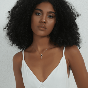 A woman with voluminous curly black hair wearing a white spaghetti-strap top and a delicate gold cross pendant necklace, photographed against a textured off-white wall.