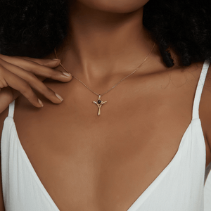 A close-up of a model wearing a delicate gold cross pendant necklace with a small dark stone at the center, styled with a white spaghetti-strap top.