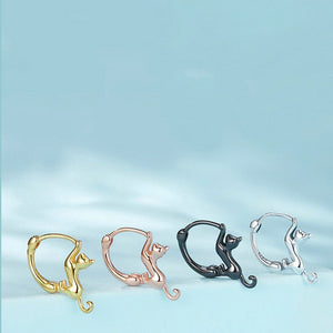 Four cat-shaped earrings in gold, rose gold, black, and silver on a light blue background.