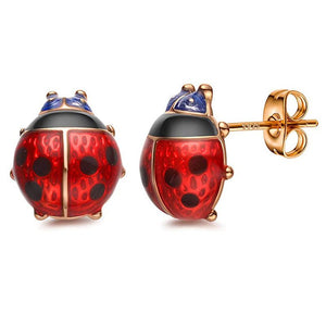 A pair of red and black ladybug stud earrings made with enamel, displayed on a white surface with a green leaf in the background.