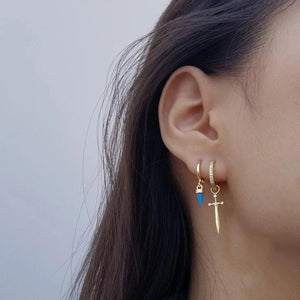 Close-up of an ear wearing gold hoop earrings with a blue gemstone and a gold sword-shaped earring.