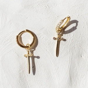 Gold hoop earrings with sword-shaped charms on a white textured background
