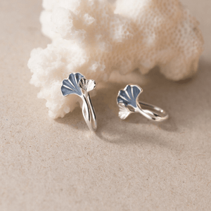 Silver ginkgo leaf earrings on a sandy surface with a shell in the background