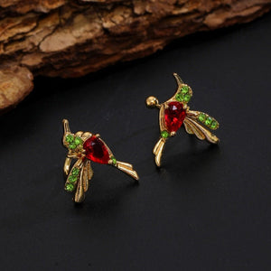 Gold hummingbird earrings with red and green gemstones on a dark surface.