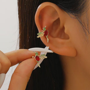 Close-up of an ear with a gold earring featuring red and green gemstones, held by a hand.