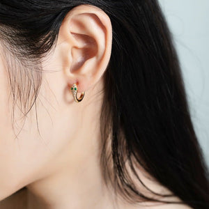 Close-up of an ear wearing a gold hoop earring with green gemstones against a neutral background