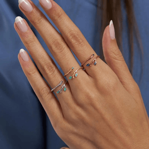 Hand wearing two delicate rings with green gemstones against a blue background