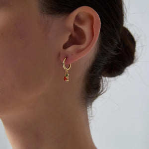 Close-up of an ear wearing a gold hoop earring with a red gemstone on a neutral background