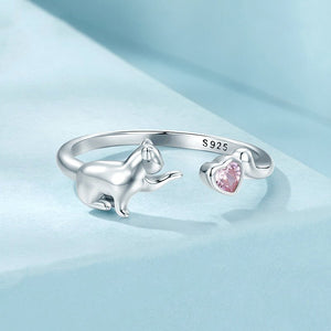 A silver ring with a design of a cat and a pink heart, adjustable in size, on a blue background.