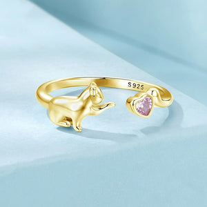 Gold ring with a cat design and pink heart-shaped gemstone on a light blue background