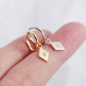 Gold hoop earrings with diamond-shaped charms on a hand against a white background