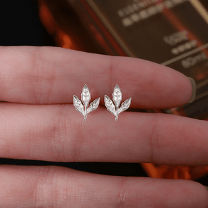 Silver leaf-shaped earrings held between fingers with a blurred background