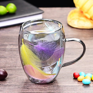 Two glass coffee cups with double wall and feather patterns, one cup is being filled with a liquid while the other remains empty, placed on a table with decorative items around.