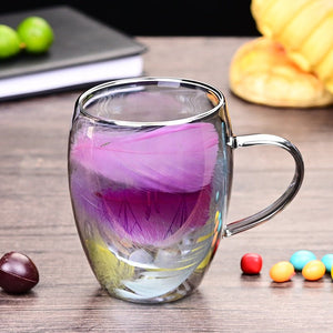 Two glass coffee cups with double wall and feather patterns, one cup is being filled with a liquid while the other remains empty, placed on a table with decorative items around.