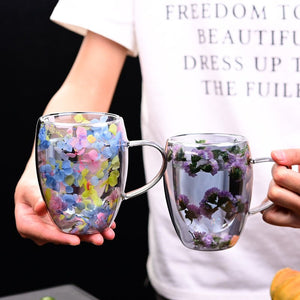 Two glass mugs, one with colorful beads and the other with flowers, held by a person wearing a white shirt.