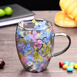Two glass coffee cups with floral patterns, one held in each hand. One cup has a pink floral design, and the other has a green and purple floral design.