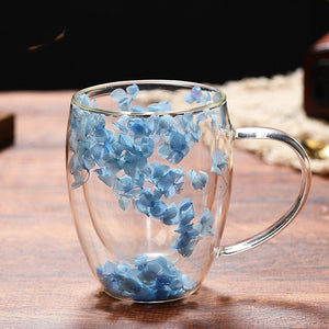 Two clear glass coffee cups with purple and blue flower petals embedded within, placed on a wooden tray.