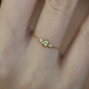 A gold-plated sterling silver stacking chain ring with a central green cubic zirconia and two smaller white cubic zirconia on the sides, displayed on a person's finger against a dark background.