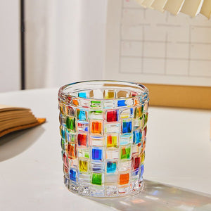 A multicolored hand-painted grid woven drinking glass with a geometric pattern, displayed on a table.