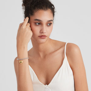 A model wearing a silver beaded bracelet with a gold heart-shaped locket charm on a light gray background