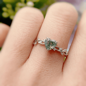 Heart-shaped green gemstone ring on a finger with a blurred natural background