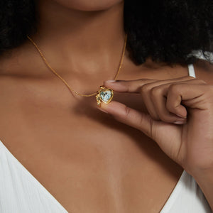 Gold heart locket necklace worn on a model, shown open to reveal a photo insert inside, held gently by hand.