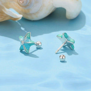Starfish-shaped earrings with turquoise and silver details on a blue surface with seashells.