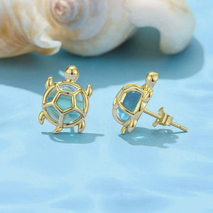Gold sea turtle-shaped earrings on a light blue surface with seashells in the background.