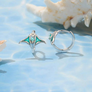 Silver manta ray hoop earrings with green gemstones on a blue textured surface with seashells.