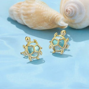 Gold sea turtle-shaped earrings with a light blue center on a light blue background with shells.