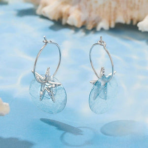 Silver hoop earrings with starfish and shell designs on a blue surface with seashells.