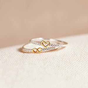 Gold and silver ring with heart and infinity symbol on a beige surface