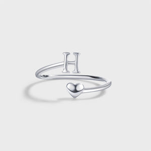 A set of three sterling silver stacking rings with bypass design, each ring with a different letter initial: R, U, and A, displayed on a white plate with dried flowers beside them.