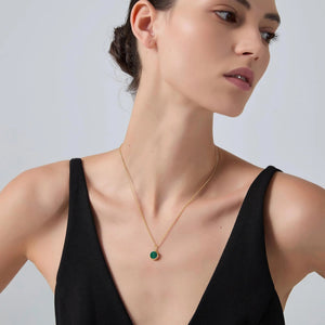 Woman wearing a gold necklace with a green pendant against a plain background