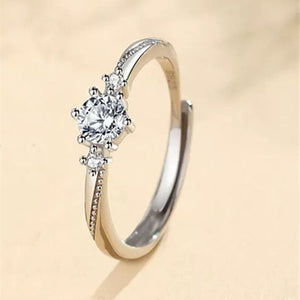 A silver wedding ring with a central cubic zirconia surrounded by smaller clear crystals, held by a shadow on a beige background.