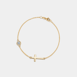 Gold bracelet with a small cross and round moissanite charm on a light gray background