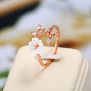 Rose gold ring with a white flower and small pink flowers on a beige stand with a blurred background