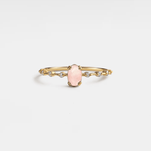 A rose quartz ring with gold plating on a silver band, featuring a single prominent rose quartz oval in the center, flanked by smaller clear stones on either side.