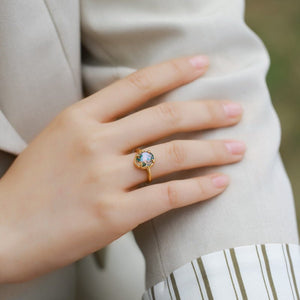 Hand wearing a gold ring with a green gemstone against a neutral background